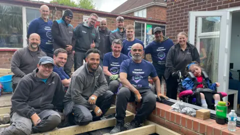 WellChild A photo of a group of men smiling into the camera. There are 12 men and they are all wearing work outfits. They are building a new garden. Also in the photo is a woman and a child in a wheelchair is next to her. Everyone is smiling. 