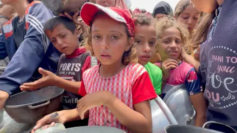 Young children wait in a line for food, looking anxious, holding empty pots in a crowded scene.