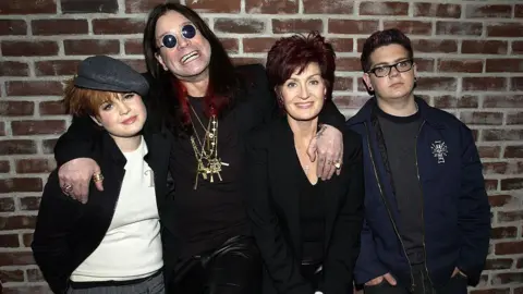 Getty Images The Osbourne family pictured in front of a brick wall. Ozzy has his arms around Kelly on his right, and Sharon, left, and all three are smiling. Jack on the far left has his hands in his pockets and is not smiling. Ozzy is wearing all-black, black round sunglasses and heavy gold necklaces. His long hair is died black and red. Kelly's short hair is dyed red and she has a grey flat cap. Sharon is also all in black. Jack is wearing glasses, a navy jacket and grey jumper.