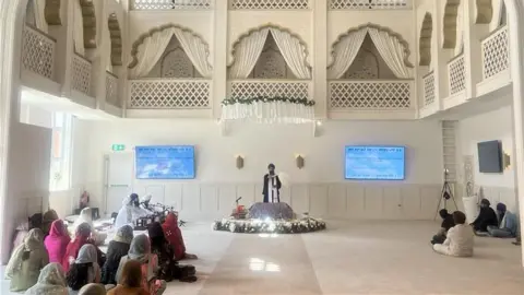 BBC/Julia Moore The main prayer room in the Gurdwara in Camberley. White room, plush carpets and arches with white and gold. Featuring men and women sitting and watching musicians and a religious leader dressed in robes with a large white feather.