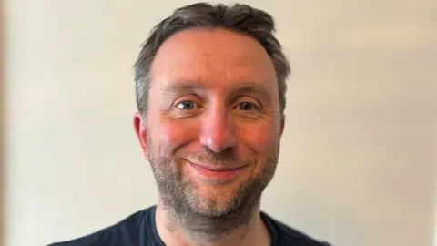 Dr James Jackson has brown and grey hair and is smiling at the camera. He is standing against a plain white background and wearing a black T shirt.