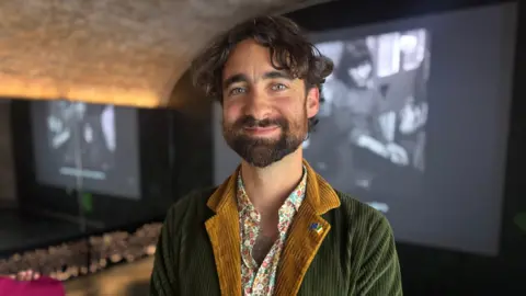 A man wearing a green jacket with black hair smiles at the camera 