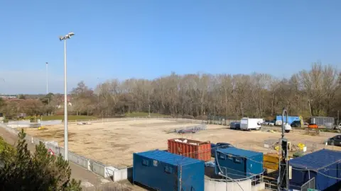 J.P. Morgan A bordered off piece of bare land. Trees are in the distance, portable buildings in the foreground.