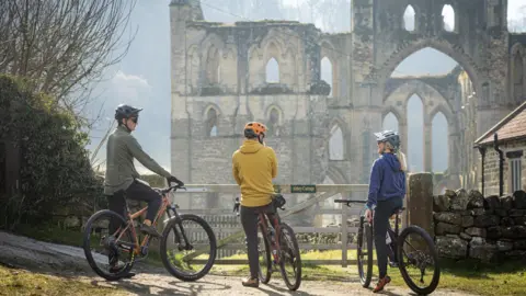 Mick Kirkman Three cyclists looking at Rievaulx Abbey from their bikes. They are stationary and standing behind a wooden gate. Two of the cyclists are men, one is wearing a green top and a black helmet, the other is wearing a mustard hoodie and an orange helmet. The other cyclist is a woman and is wearing a blue hoodie and a grey helmet with a blonde ponytail sticking out of it.