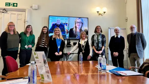 Handout Seven women and one man stand in front of a table with a television behind them. They are all looking at the camera and smiling. One lady is wearing a chain of office around her neck and some are wearing lanyards. There's some paperwork and water bottles on the table. The television screen is split in two, with a woman on one side and a man on the other. 