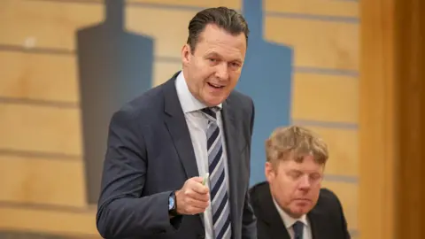 PA Media Russell Findlay has short brown hair and wears a grey suits while standing and addressing the Holyrood chamber. He points a pen in front of him. There is a man behind him with short fair hair who is sitting down and blinking