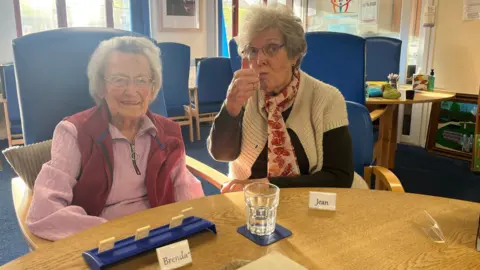 Two women sat a table in a day care centre. One is wearing a pink top with dark pink sleeveless jacket. The second woman has a black top, white jacket and red and pink scarf. Both women are wearing glasses