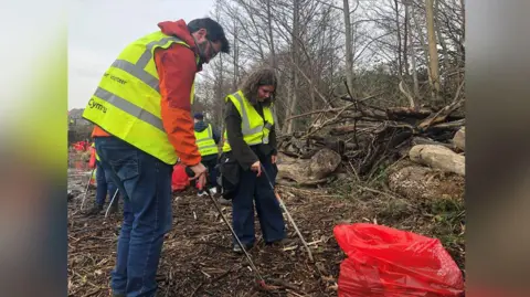 Two volunteers picking up litter on the bank of the River Taff. A red bag lies at their feet. One volunteer is wearing blue skinny jeans, an orange jacket and a yellow high-vis jacket. Another volunteer stands slightly behind him, she is wearing a black jumper and trousers, a yellow high-vis jacket and she has curly brown hair. Trees are visible in the background. 