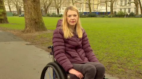 Chair of the Spinal Research charity Tara Stewart who was paralysed from the chest down herself in a similar horse riding accident. She is sitting in her wheelchair on a path in a wooded area. Ms Stewart has a purple hooded coat on and has long blonde hair and black jeans.