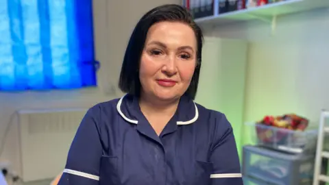 BBC Kelly Harwood has black hair worn in a bob. She is wearing a navy blue nurse's uniform and is looking at the camera. There are medical supplies and folders on shelves behind her.