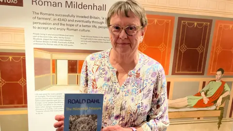 Vikki Irwin/BBC A head and shoulders image of Kay Finnis. She is standing in a museum and holding a blue book which reads Roald Dahl The Mildenhall Treasure. She is wearing glasses and a colourful patterned top and looking into the camera. 