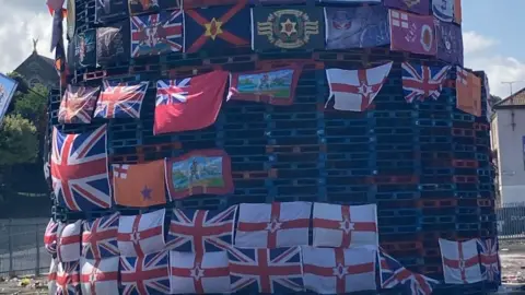 Shows a large amount of union flags and other emblems on a large unlit bonfire
