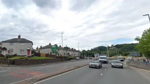 Google A dual carriageway with a junction with a smaller road on the left. There is a roundabout and a tree-covered hill in the background.