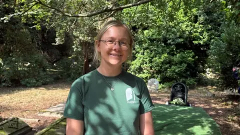 A woman with tied back blond hair wears a green t-shirt and round glasses. She is standing in a cemetary. Behind her a black marble gravestone can be seen with white flowers laid in front of it.