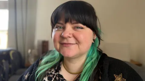 Yolanda Shirin/BBC A close-up shot of a woman with a black fringe and teal coloured hair. The lady has several nose piercings and is wearing black. 