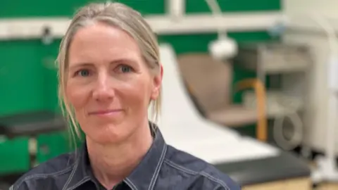 Dr Nicki Mazey sits in a doctor's surgery with an examination bed behind her. She has blonde hair which is tied back and wears a dark denim shirt and smiles at the camera.