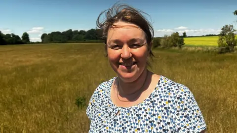 Prof Cannon stood in a spotty top in front of a field full of grass. The grass is yellowy brown.
