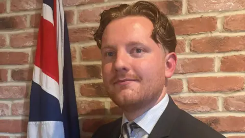 Reform UK's Chris Eynon stands in front of a brick wall and Union Jack flag wearing a black suit, striped tie and blue checked shirt.