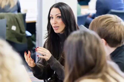 A woman with long black hair, wearing a brown jumper and with red nail varnish is speaking in a group of people 