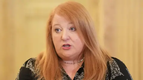 PA Naomi Long has shoulder-length red hair and is wearing a black and white blouse.