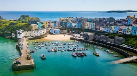 Around 80 small boats moored in green-blue water in the harbour in Tenby, viewed from above in bright sunshine. There are rows of multi-coloured houses behind the harbour - green, blue, yellow, turquoise, orange and pink - and a view out to sea at the top of the frame.