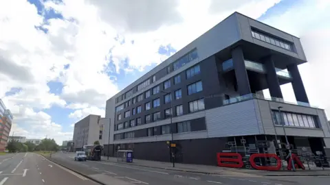 Image taken from the road outside of Birmingham Ormiston Academy (BOA). Large red letters are seen at the base of the building reading BOA. The building itself is at least six storeys high and is dark grey and light grey in colour. 
