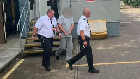 A man with a blurred face being led down stairs from a portable cabin by security officers