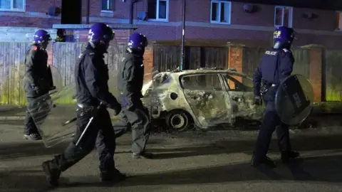 PA Media Four police officers wearing riot gear walking past a burnt-out car.