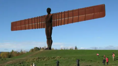 Geograph/Adrian Taylor The Angel of the North, a very tall copper/brown statue with large, wide ridged wings and a ridged body. It is on a green, grassy hill, and there are some people gathered around it. The blue sky is visible in the background, with a few clouds.