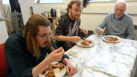 Three men of different ages sitting down together at a table, all eating pie. 