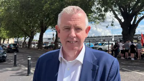 John Watson standing in front of a picket line wearing a suit and a shirt