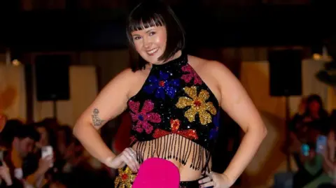 A woman wearing a glittery black dress, with a glittery pink stoma bag, smiles.