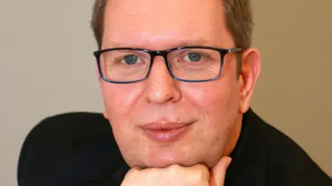 Wombourne Choral Society A close-up head shot of a man in a black jacket who has brown hair and is wearing glasses. 