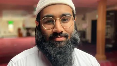 A man with a black and gold round glasses. He is wearing a white shirt and white hat. He is stood inside a mosque and is looking into the camera.