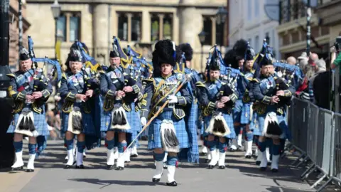 A military band wearing blue checked kilts, black bearskin hats and carrying bagpipes marches through a city. 