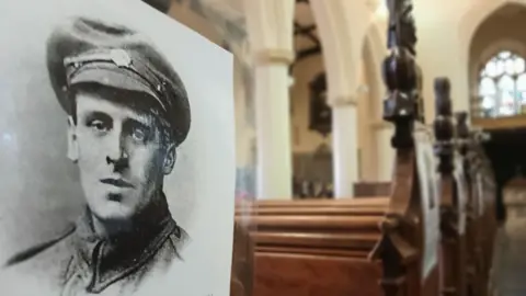 Ipswich War Memorial Project A close up of a portrait of a serviceman from World War One. It has been put in place at the end of a church pew. Further church pews can be seen behind it along with a stained glass window. 