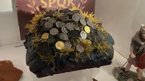 A collection of coins sat on a pretend rock covered in moss in a display cabinet at the museum