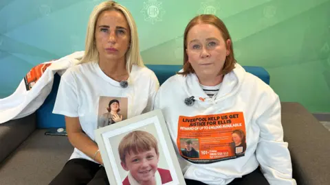 Ellis's mum Carolyn Cox (left) is a blonde woman. She sits next to Ellis's auntie Julie O'Toole (right) who has ginger hair. Both wear hoodies and t-shirts showing Ellis's face and hold a photograph of Ellis as a child.