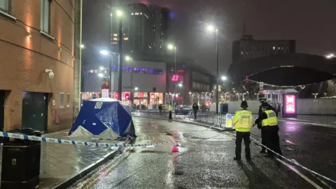 BBC A wet road at night taped off by white and blue police tape. Police officers in yellow hi vis jackets and black hats are standing next to it. There is a blue and white West Midlands police tent on the pavement on the left.
