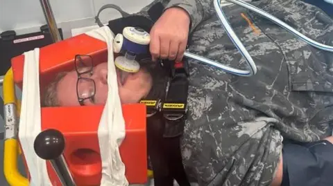 Josh Howell A man laying down with a neck brace on. He has a tube in his mouth.