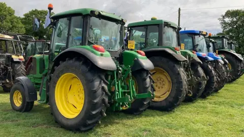 Tractors are lined up in a field, ready to take part in the convoy. There are several green tractors and there is one blue one.