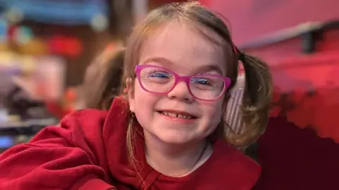 Tilly smiling at the camera she has brown hair in ponytails and is wearing pink glasses and a red dress