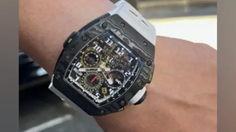Metropolitan Police Close-up of a person wearing a luxury Richard Mille wristwatch with a black carbon-effect tonneau case, complex exposed movement dial, and a white strap.