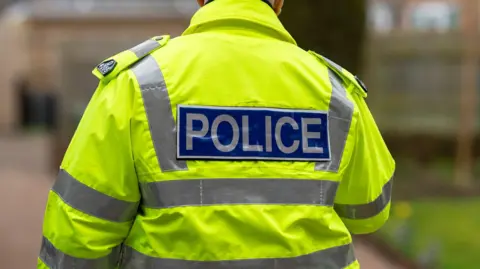 A police officer seen on a residential street from behind. He is wearing a high-vis jacket with the words "police" on