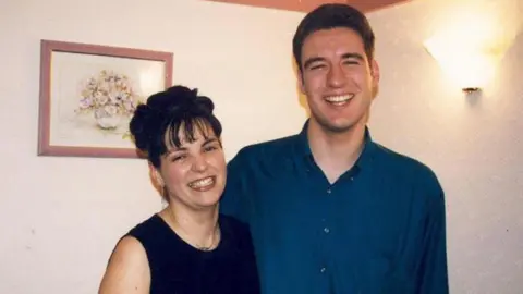 Mr and Mrs Wilson are standing side by side in a room. They are smiling. Behind them is a painting of a vase and flowers and a light.