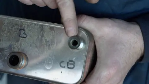 A man holding a silver metal piece of appliance from a boiler, pointing to a hole that has become clogged.