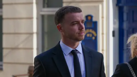 Lewis Carr walks towards Jersey's Royal Court. He is wearing a dark suit with a white shirt and dark tie. He has a neatly trimmed beard.