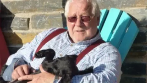 Tom Marshall A man with grey hair and red glasses holding a small black dog 