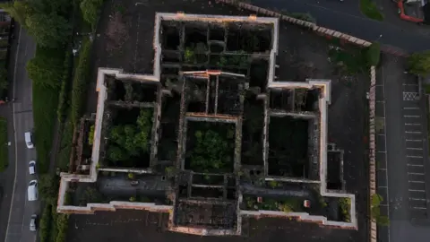 An aerial drone image of Crumlin Road Courthouse which shows the inside
of the structure which has lost its roof. Vegetation is growing up through several of the rooms