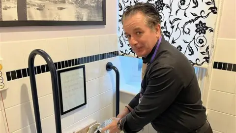 Phil Harrison/BBC A man in a black jumper is pouring water in a toilet tank.